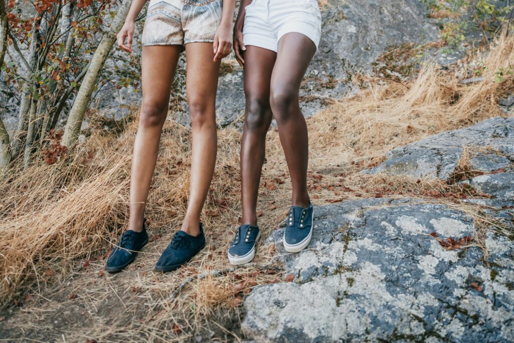 Close-up of two women's legs in shorts and shoes standing on rocky landscape with dry grass.